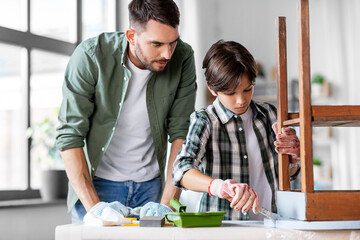 renovation, diy and home improvement concept - father and son in gloves with paint brush painting old wooden table in grey color