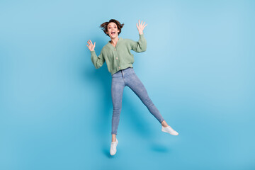 Fototapeta na wymiar Full length photo of cute impressed young lady wear green cardigan jumping high isolated blue color background