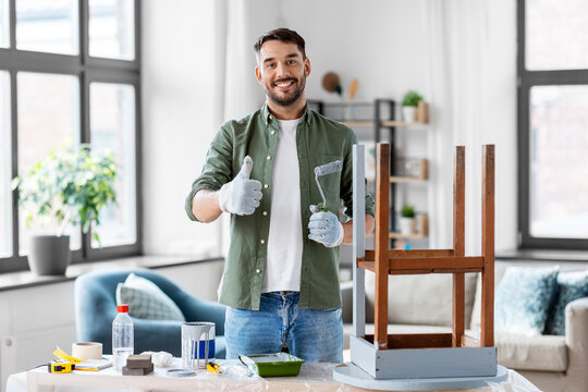 renovation, diy and home improvement concept - happy smiling man in gloves with paint roller painting old wooden table in grey color and showing thumbs up