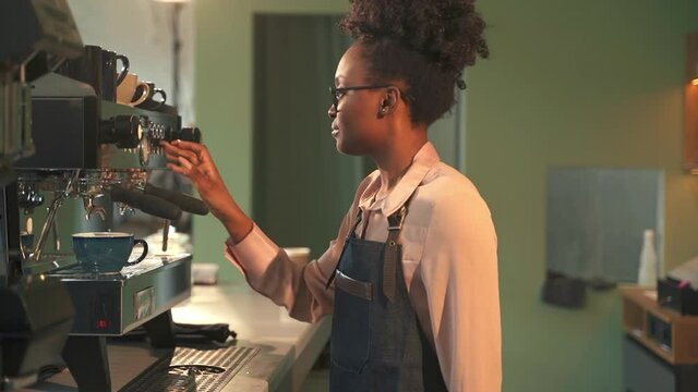Barista black lady works on coffeemaker. Spbas Happy African-American lady with curly hair prepares coffee with automatic equipment at counter in cozy cafe