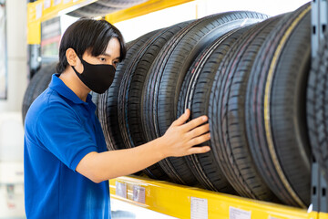 Fototapeta na wymiar Asian male worker wear face mask checking new tire wheel on shelves shelf at wheel store. Salesman examining new tire at workshop. Car service and Maintenance concept