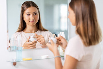 beauty, hygiene and people concept - teenage girl with moisturizing cream looking in mirror at bathroom