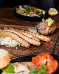 Stack of cooked sausages served on a rustic wooden board with sauce and baked potatoes.
