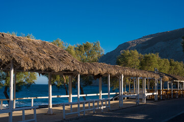 Black sand beach on Santorini during covid-19 lockdown. Empty street. Green trees on shore. Huge rock. Nobody. Straw umbrellas.