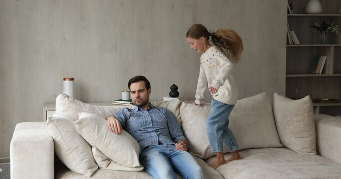Young tired father sit on sofa at home feels exhausted by noisy little daughter makes noise jumping near irritated annoyed dad. Parent and difficult hyperactive misbehaved child need attention concept