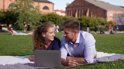 Young couple lying on lawn and watching laptop content on beautiful sunny day.