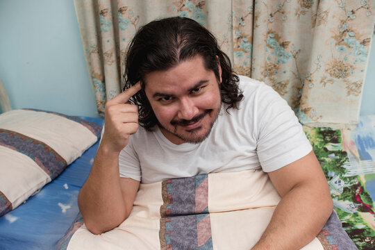 A lazy and smart aleck bearded guy in his 30s thinks he is clever pointing to his brain while sitting on the bed after a nap.