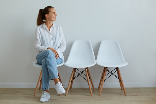 Indoor portrait of young attractive woman sitting on chair in waiting room, wearing jeans and white t shirt, looking away with charming smile, expressing positive emotions.
