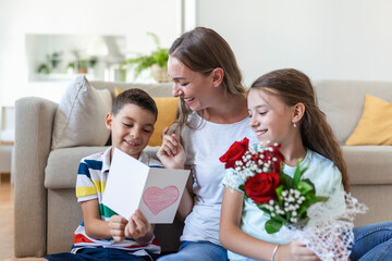 Young mother with a bouquet of roses laughs, hugging her son, and cheerful girl with a card congratulates mom during holiday celebration in kitchen at home