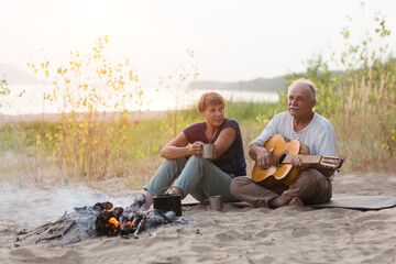 Senior couple Camping holiday in the summer nature. Camping concept. senior man playing guitar and singing song to his wife sitting on the beach near the fire
