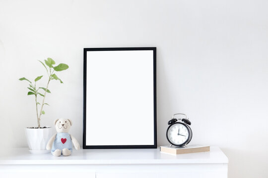 Interior poster mock up with vertical black frame, green plant in a pot, bear toy, book and clock on a white table and white wall background, scandinavian style