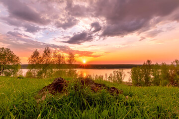 Scenic view at beautiful sunset on a shiny lake with old rough stone on the foreground, green grass, birch trees, golden sun rays, calm water ,nice cloudy sky on a background, spring landscape