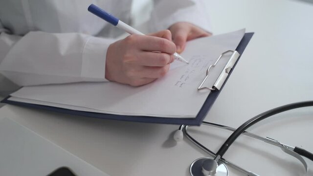 Doctor medical care work. Spbas Young internist in uniform writes symptoms and complaints on clipboard sitting at workplace in light hospital office closeup