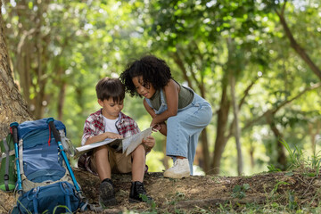 African American ethnicity boy and girl children wearing plaid shirt and backpack sitting at tree base talking and looking down on the map travel on hand in the park 