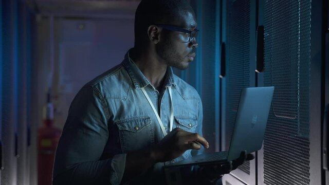 Male programmer working with laptop and standing in dark interior of render farm spbas. Close-up view of young African man types text and holds computer in hand, examines equipment and stands in room