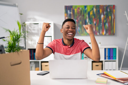 Ambitious hardworking handsome man of African American descent sitting in office in front of computer got good news, won a tender, got a promotion, happy raises hands in air, win, victory