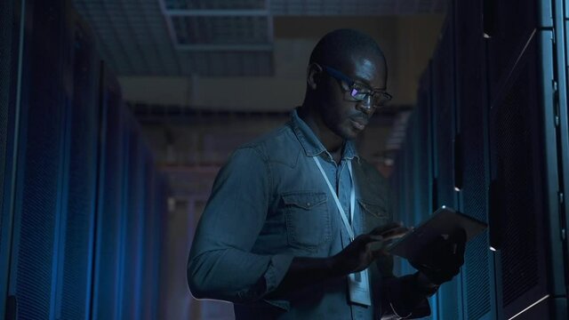 Male engineer using tablet and examining networks while standing in data center spbas. Close-up view of young African man holds device in his hand and inspects digital equipment racks, stands in dark