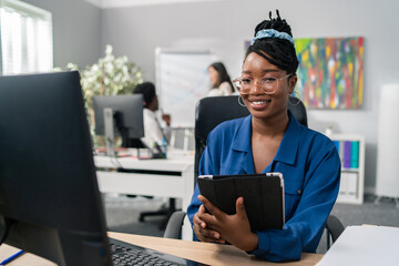 Elegant businesswoman dressed in a neat blue shirt and glasses sits in the office at the desk, in hands holding a tablet, smiling at camera, busy morning at company, co-workers in background