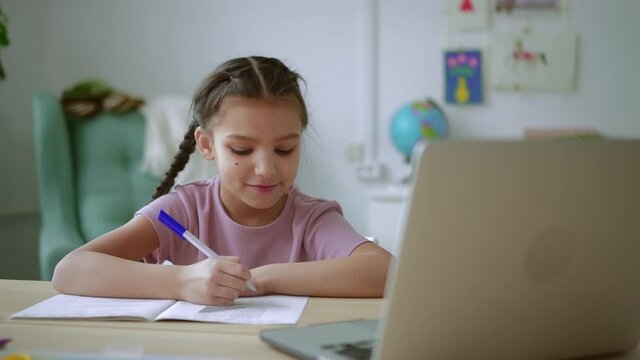 Home education of junior schoolchildren. Spbd Smiling girl student writes in copybook listening to tutor during video lesson via laptop at desk in room