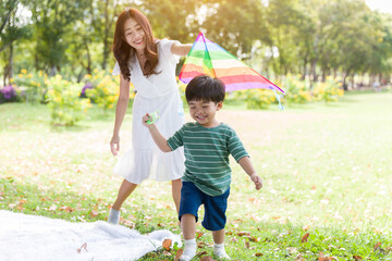 Happy cheerful Asian little boy running and playing with kite flying with mother outdoor in the park. Little boy and mother having fun, enjoy, relaxing with kite flying together. Family, kid, playing