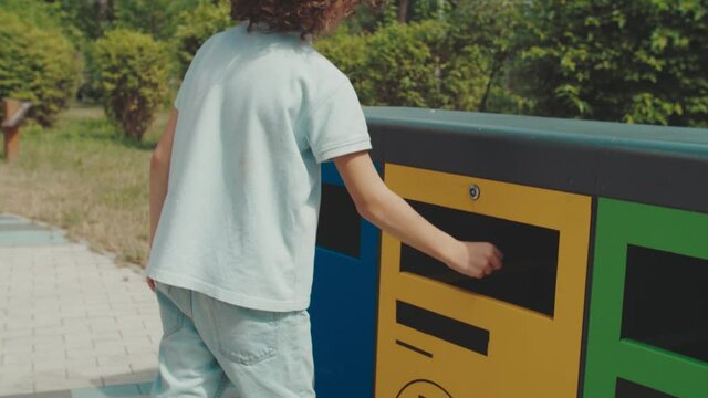 Curly boy throwing away empty plastic bottle into recycle dustbin outdoors. Environmental aware school kid using correct garbage bin because of eco-education and conversance of sorting waste