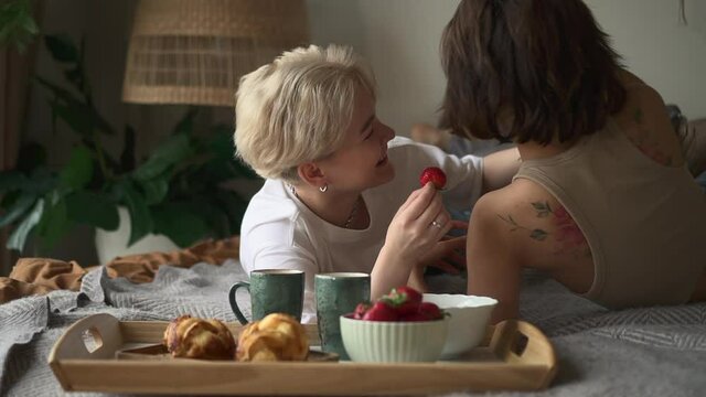Lesbian couple romantic relationship. Spbd Blonde lady teases Asian friend feeding with strawberry and kisses in bedroom at home in lazy morning