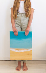 Female artist holding a resin art painting with blue ocean waves and beach on wood background.