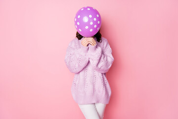 Portrait of young girl holding in hand air ball closing face wearing oversize isolated over pink pastel color background