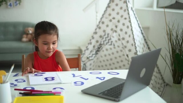 Online primary school education. Spbd Pretty little Korean girl student answers teacher question to laptop screen at table with digits in room