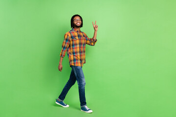 Photo of pretty sweet dark skin guy dressed checkered shirt glasses walking showing v-sign empty space isolated green color background