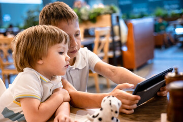 Boy and toddler playing video games on tablet online on internet while waiting orders in restaurant. Children using smartphone. Kids using technology. Screen time addiction.