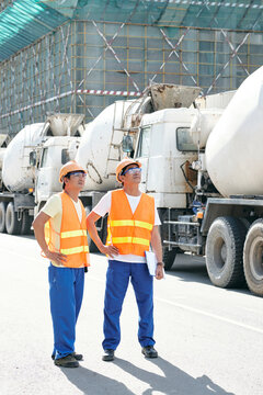 Smiling contractors walking along rows of concrete mixer trucks and planning working day at construction site