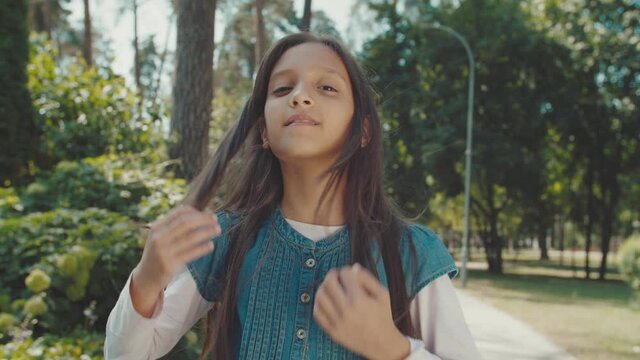 Portrait of beautiful middle eastern girl with backpack in casual clothing combing her straight hair, looking directly into camera, standing in public park at morning.