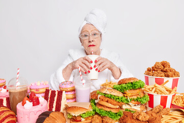 Surprised elderly lady with red lipstick has imbalancd nutrition eats different tasty junk food drinks cocktail containing much sugar dressed in domestic clothes. Old people and cheat meal concept