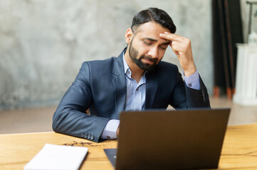 Upset Indian businessman in formal suit has strong headache, sitting in front of laptop and holding...
