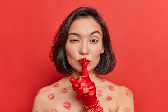 Mysterious Asian woman with dark hair makes silence gesture tells secret information poses shirtless wears red lipstick makes shh sound looks self confident at camera. People and secrecy concept