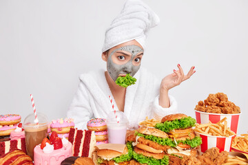 Displeased Asian woman in bathrobe looks with indignant expression has mouth full of green salad keeps to diet for weight lose eats healthy food to boost immunse system applies beauty mask on face