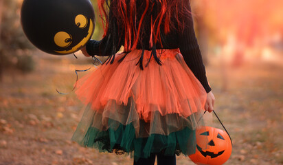 Little girl in witch costume with a balloon and a pumpkin bucket playing in autumn park. Child...