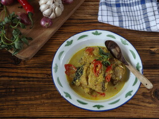 Mangut pe or spicy fish curry is Javanese cuisine. Smoked stingray fish cooked with spices and coconut milk. Served in enamel plate with ingredients around. Rustic wooden background.
