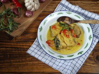 Mangut pe or spicy fish curry is Javanese cuisine. Smoked stingray fish cooked with spices and coconut milk. Served in enamel plate with ingredients around. Rustic wooden background.