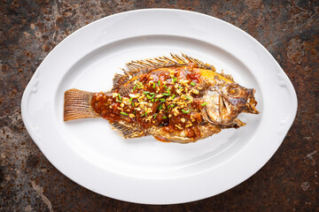 tasty large deedp fried nile tilapia fish with chlli sauce, fried garlic and sliced spring onion in oval ceramic plate on rusty texture background, top view