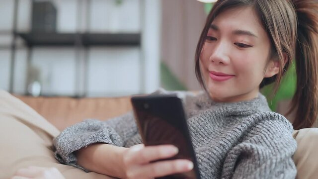 social media enjoy,asian female adult smile laugh hand seach surf smartphone device social media addict at sofa living room home interior background,home quarantine concept