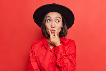 Pretty Asian girlfriend keeps hand near rounded lips makes mwah wears stylish headwear and shirt has romantic mood prepares for date poses against bright red background. Human face expressions