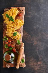 Tagliatelle. Homemade pasta, basil leaves, flour, pepper, olive oil, cherry tomato and rolling pin and pasta knife on dark old wooden background. Food concept. Mock up. Horizontal with copy space.