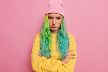 Studio shot of offended displeased hipster girl with long dyed hair keeps arms folded doesnt speak to boyfriend after quarrel feels angry has bad mood wears yellow jumper and hat. Negative emotions