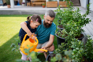 Father and small daughter with watering can outdoors in tha backyard, looking at camera.