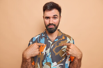 Serious bearded man points at himself asks who me has attentie look at camera wears coloful shirt...