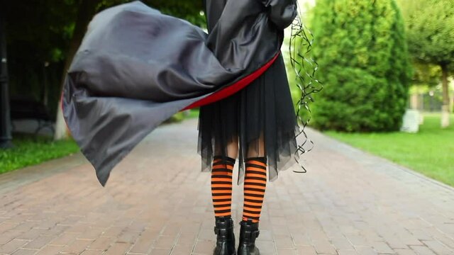 Teen girl in halloween costume like a witch with orange stripped socks and long black cloak playing with her clothes on the street. Holiday concept. Slow motion.