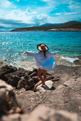 Beautiful woman seen from behind standing on a stone beach on the shore of Vis island in Croatia. Waving her arms hapily while posing, standing on one leg and meditating in amazing summer weather