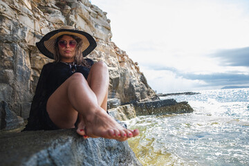 Attractive girl wearing a black dress, sunglasses and a hay hat sitting by the sea. Stormy day by the beach in Croatia, warm summer of 2021. Barefoot feet in foreground